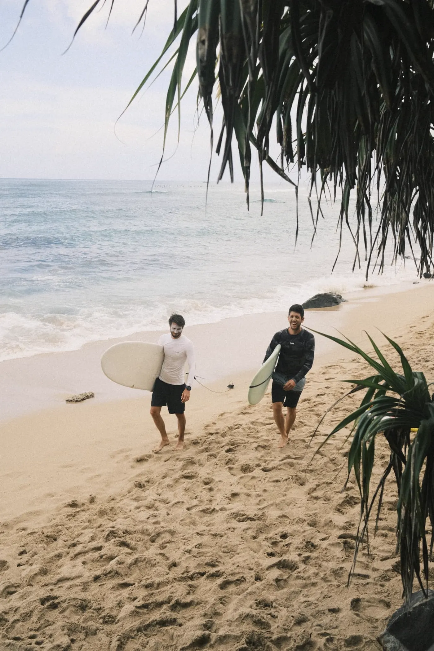 Két surfer sétál a parton deszkával